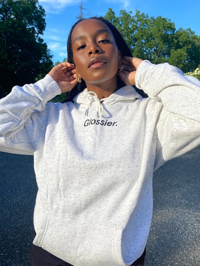 A woman with long dark hair wearing a white hoodie with the text "Glossier."
