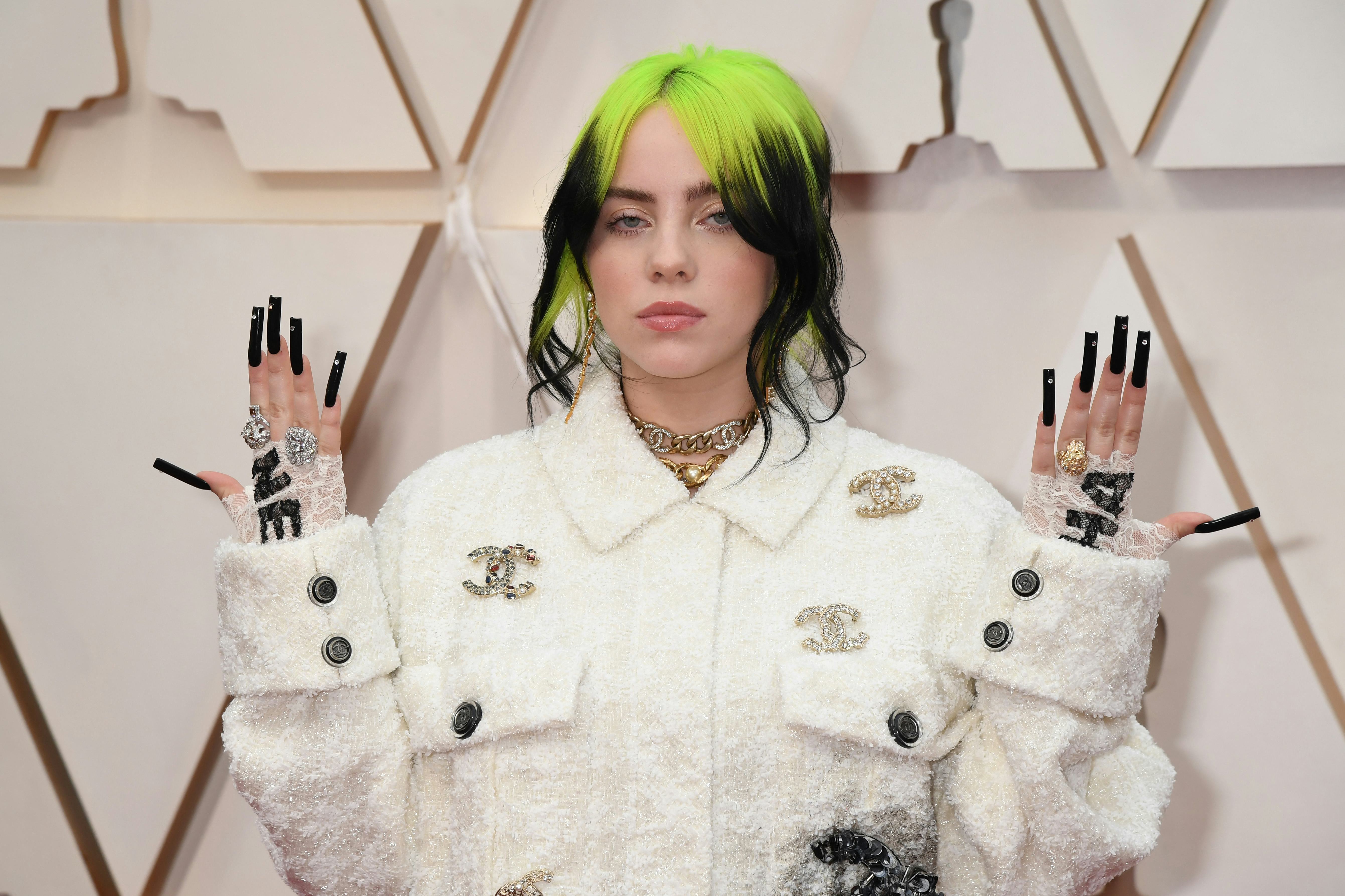 Billie Eilish, with black and green ponytail, holds hands up, showing off black manicure