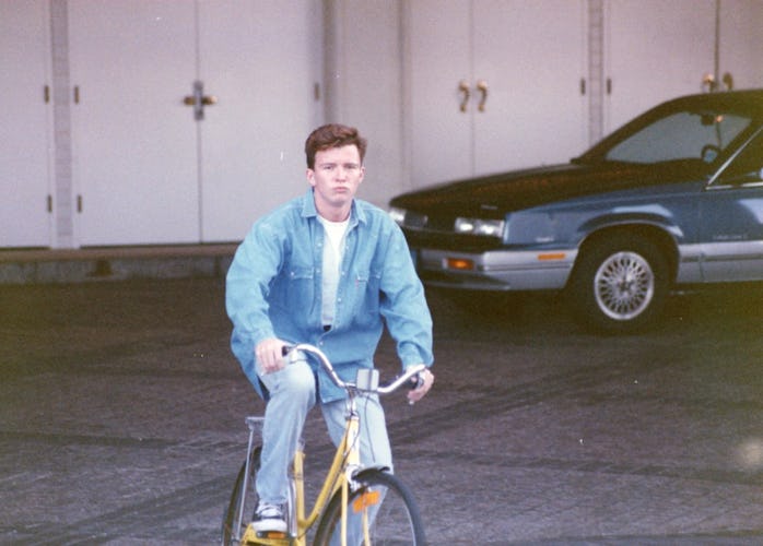 A young Rick Astley can be seen on a bicycle in Vegas. There is a car parked behind him.