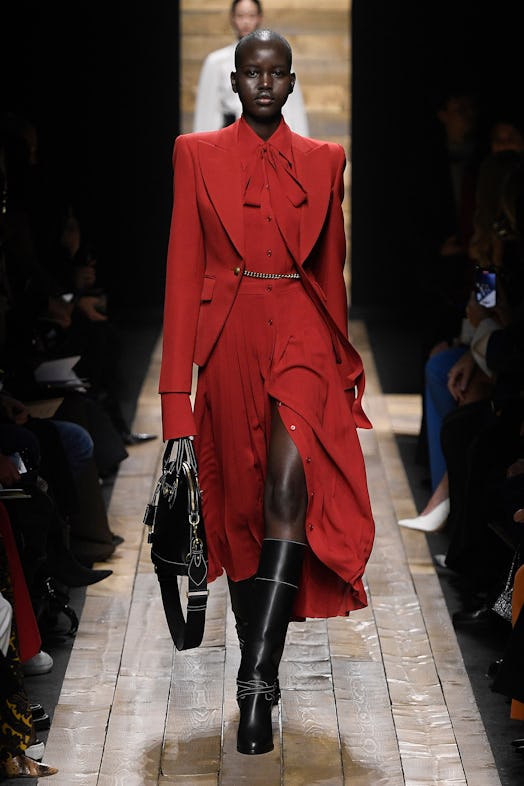 The model wears a red button-down dress with a bow tie, a matching red blazer, and knee-high black b...