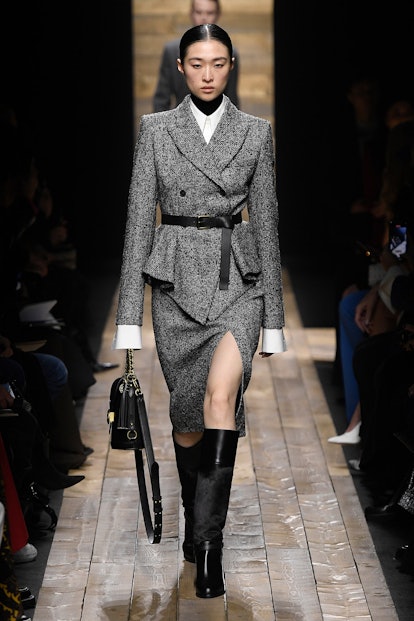 The model wears a grey belted blazer, a matching skirt with a front slit, and knee-high black boots ...