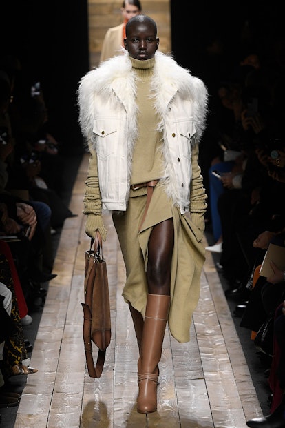 The model wears a beige turtleneck, a matching skirt with a front slit, and a white fur vest by Mich...
