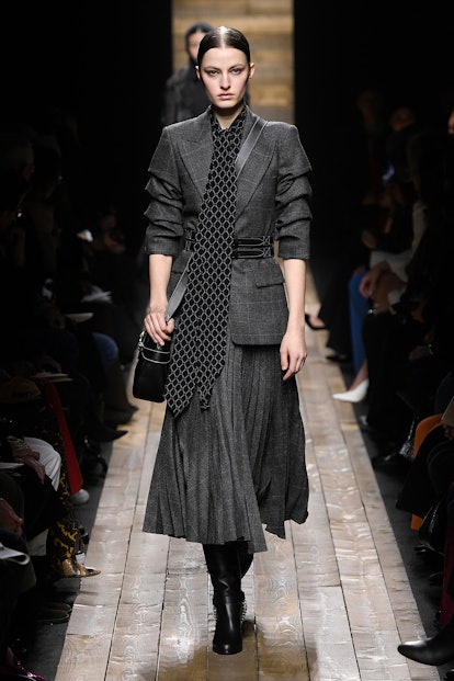 The model walks the runway in a grey checkered blazer, matched with a pleated midi skirt and a long ...