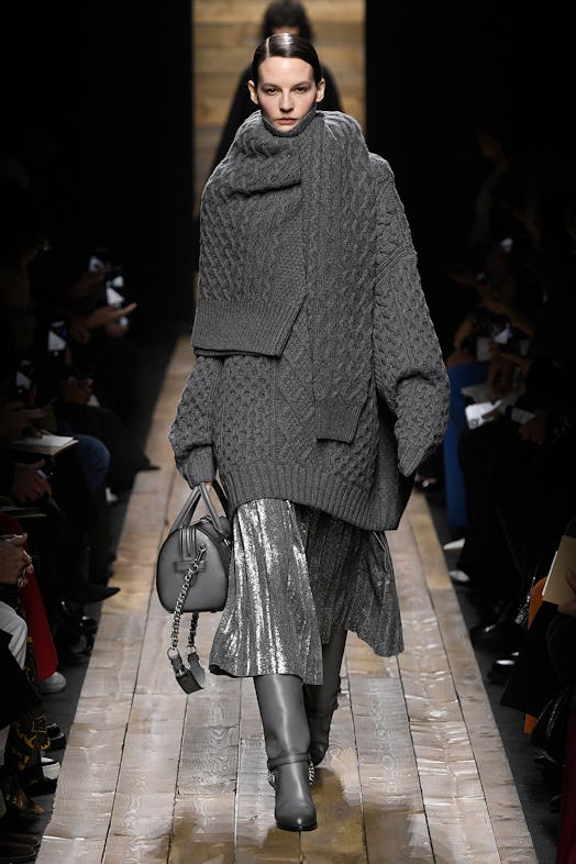 A model walks the runway in a grey cable-knit sweater, matching cowl and a metallic-grey pleated ski...