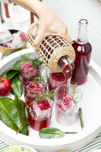 Pomegranate juice is poured from a wicker bottle over ice cubes containing its seed and rosemary gar...