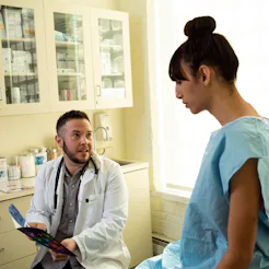 A transgender woman in a hospital gown speaking to her doctor, a transgender man, in an exam room. C...