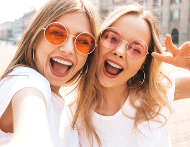Two women wearing circular-shaped sunglasses and white T-shirts smile and snap a selfie.