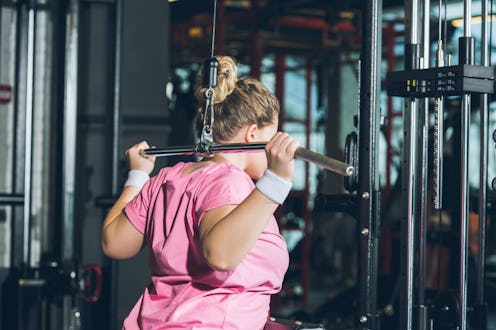 A person wearing a pink t-shirt and white wrist bands performs a lat pulldown behind her head. Gym a...