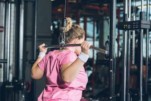 A person wearing a pink t-shirt and white wrist bands performs a lat pulldown behind her head. Gym a...