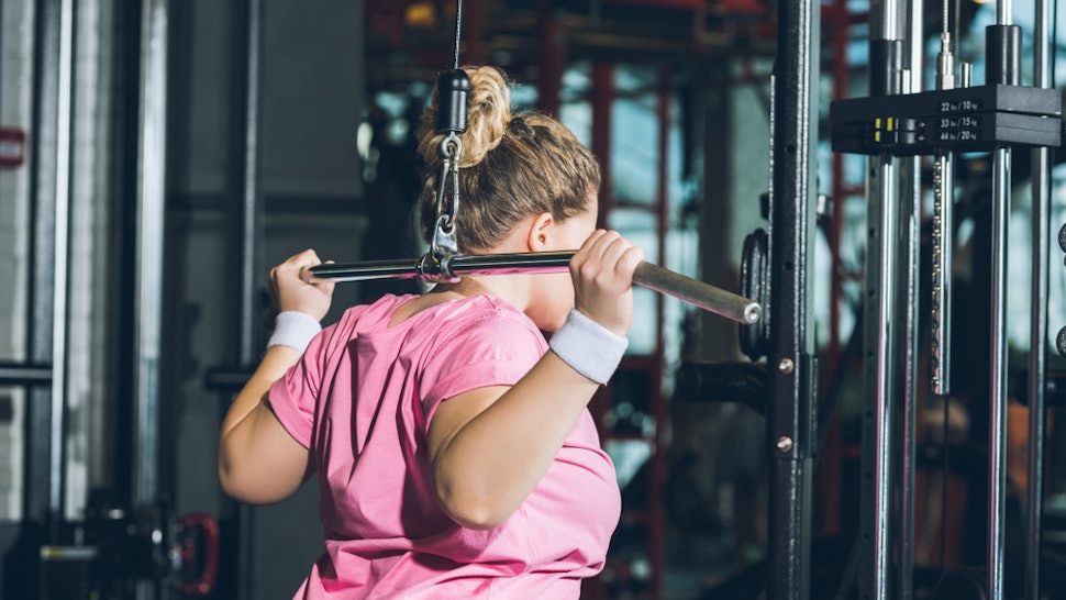 A person wearing a pink t-shirt and white wrist bands performs a lat pulldown behind her head. Gym anxiety may be keeping you from feeling comfortable in the gym, but there are ways to overcome it so you can have a great workout.