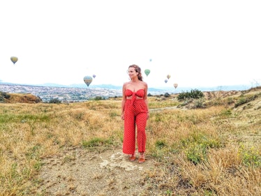 A woman in a bright red jumpsuit smiles and looks off to the side in a field with hot air balloons i...
