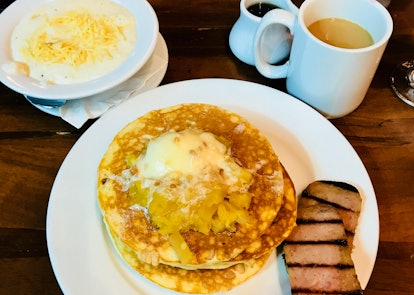 A plate of macadamia nut pancakes with pineapple and spiced ham is placed next to Kona coffee and gr...