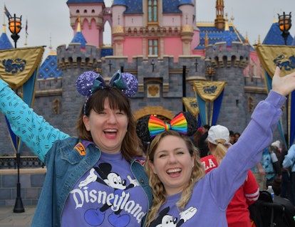 Two friends wearing matching purple Disneyland sweaters and Mickey ears stand in front of the castle...