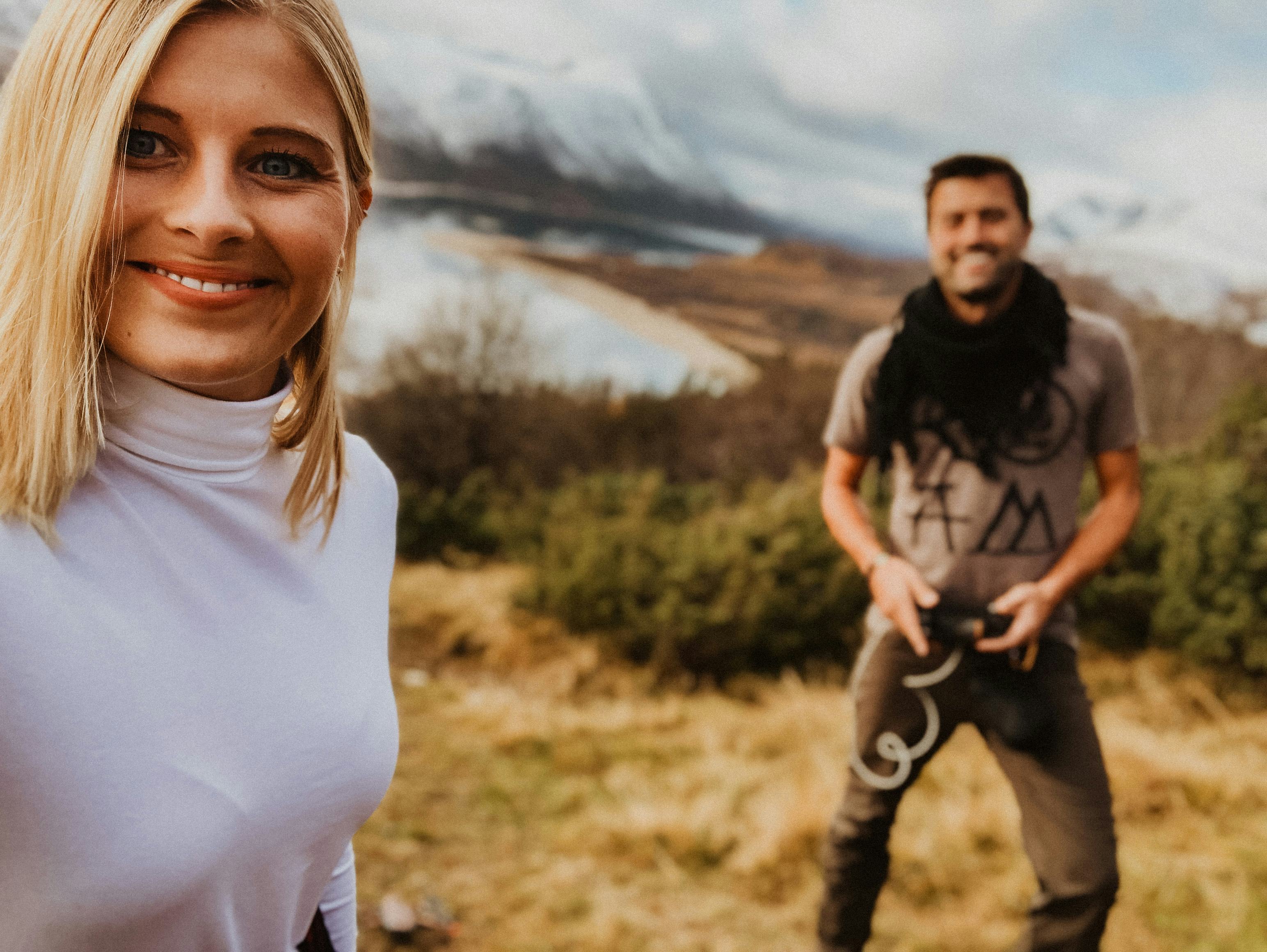 A blonde woman smiles while taking a selfie in a field with mountains in the background on a sunny d...