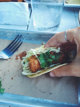 A woman holds a fish taco in her hand over a small, metal tray.