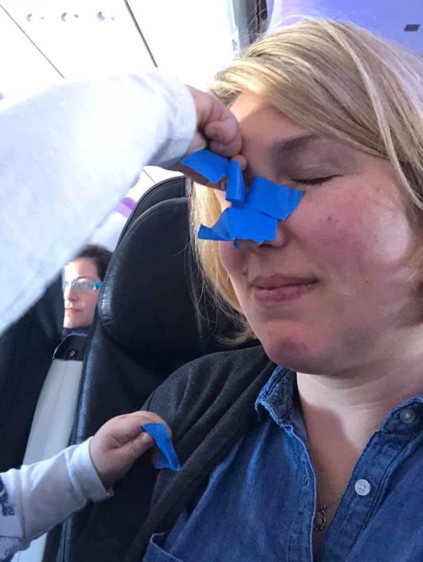 Mom on a plane with blue tape all over her face with toddler hand putting it on