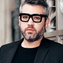 A portrait of Brandon Maxwell in a black suit and black glasses