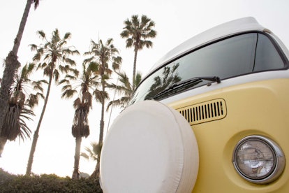 A 1971 Volkswagen Westfalia is yellow and very vintage-looking against a sky filled with palm trees.