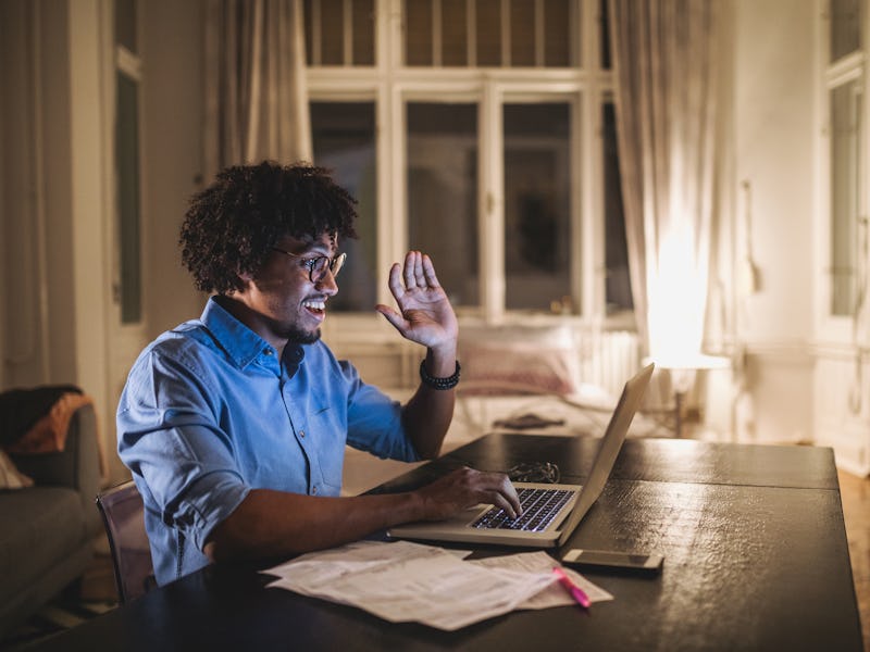 A curly-haired man in a blue shirt waving at someone during a video call and smiling