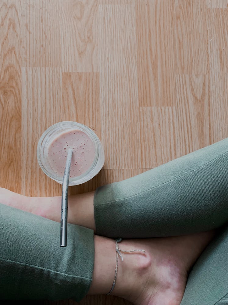 A young woman sits on a hardwood floor with a smoothie after working out at home.