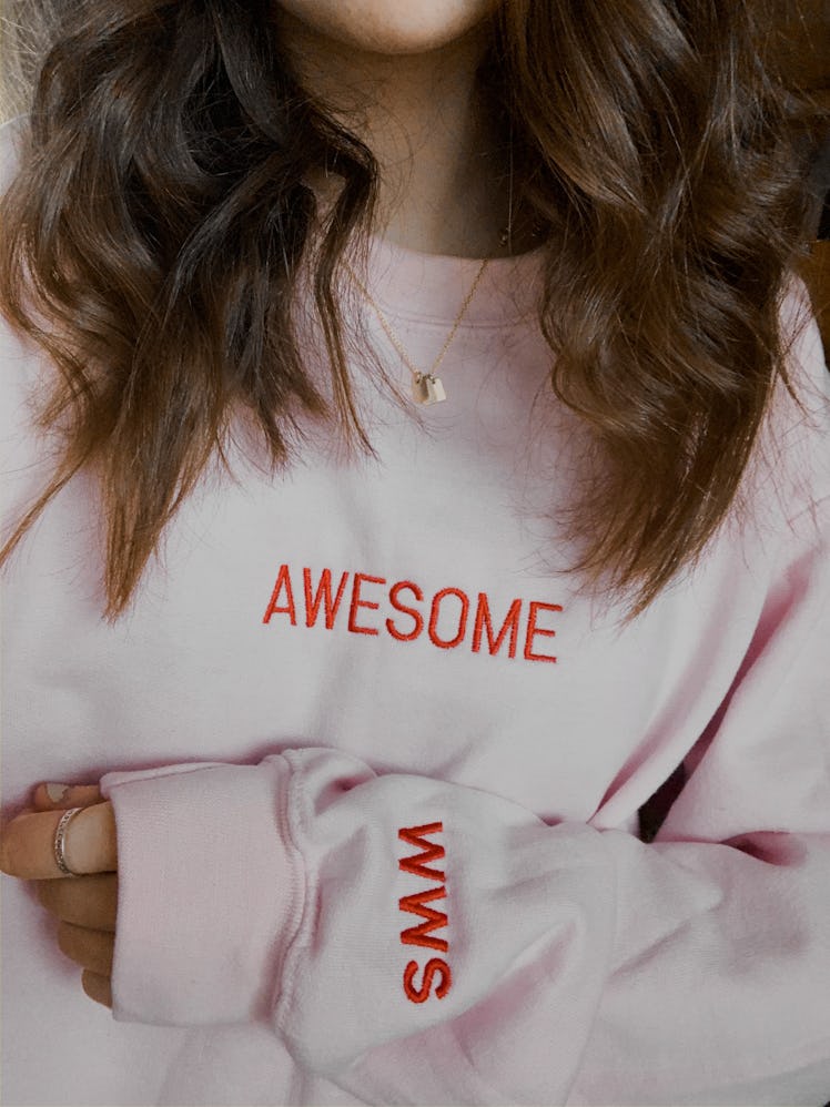 A young woman poses in a light pink sweatshirt that says, "Awesome."