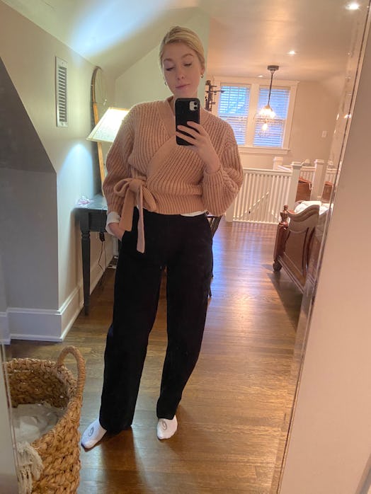 A young woman taking a photo of herself in the mirror in a light brown sweater, black pants, and whi...