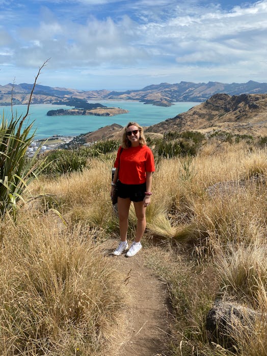 A blonde woman wearing sunglasses, black shorts, and a red tee smiles and stands on a hilltop with t...