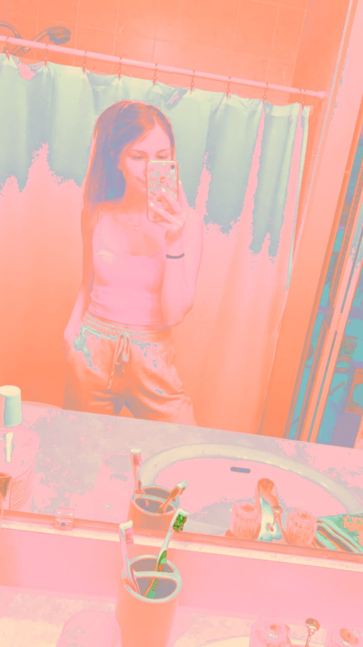 A young woman poses in a bathroom mirror for a selfie with a colorful Instagram filter.