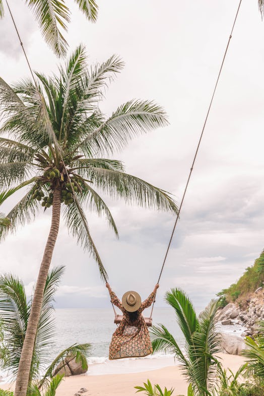A woman in a flowy dress swings on a long rope swing in the middle of palm trees
