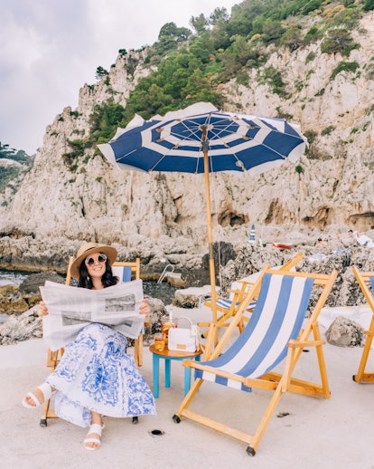 A woman sits on a striped beach chair while reading newspaper on a beach in Capri.