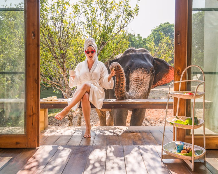 A woman in a bathrobe and towel around her hair sits on a porch with an elephant next to her.