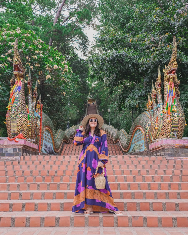 A woman in a colorful maxi dress and big sun hat poses on temple stairs in Thailand.