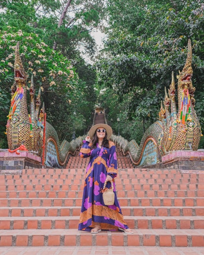 A woman in a colorful maxi dress and big sun hat poses on temple stairs in Thailand.