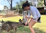 A woman smiles and bends down to feed a kangaroo.