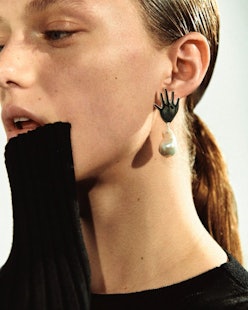 A model holding her hand to her mouth while posing with earrings in the shape of a hand