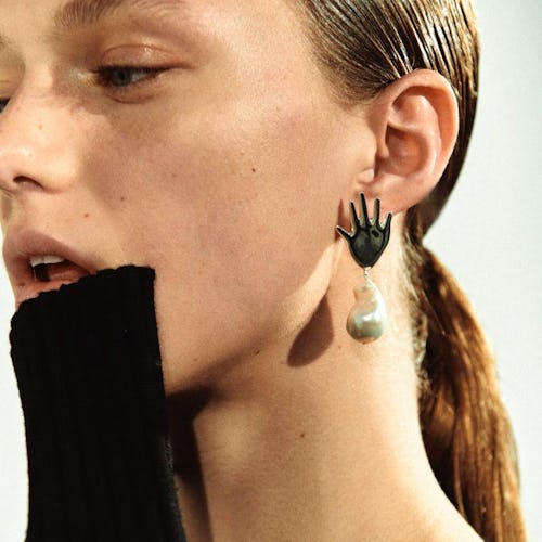 A model holding her hand to her mouth while posing with earrings in the shape of a hand