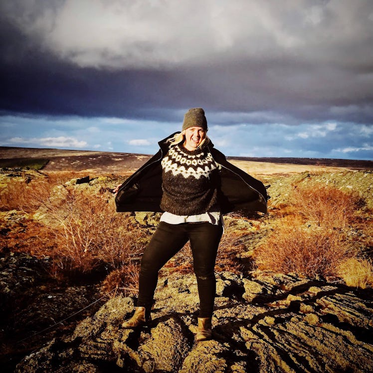 A woman wearing a beanie and winter sweater poses on a pile of rocks in a field on a sunny day.