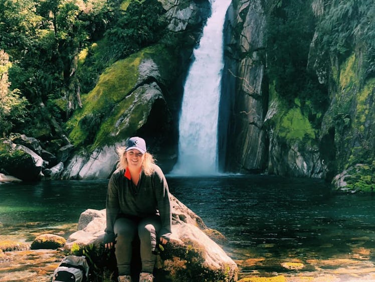 A woman wearing a baseball hat and athletic apparel sits on a rock near a body of water.