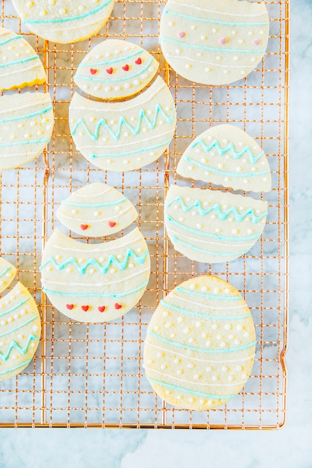 Several sugar cookies cut into egg shapes with decorative frosting on a copper cooling rack
