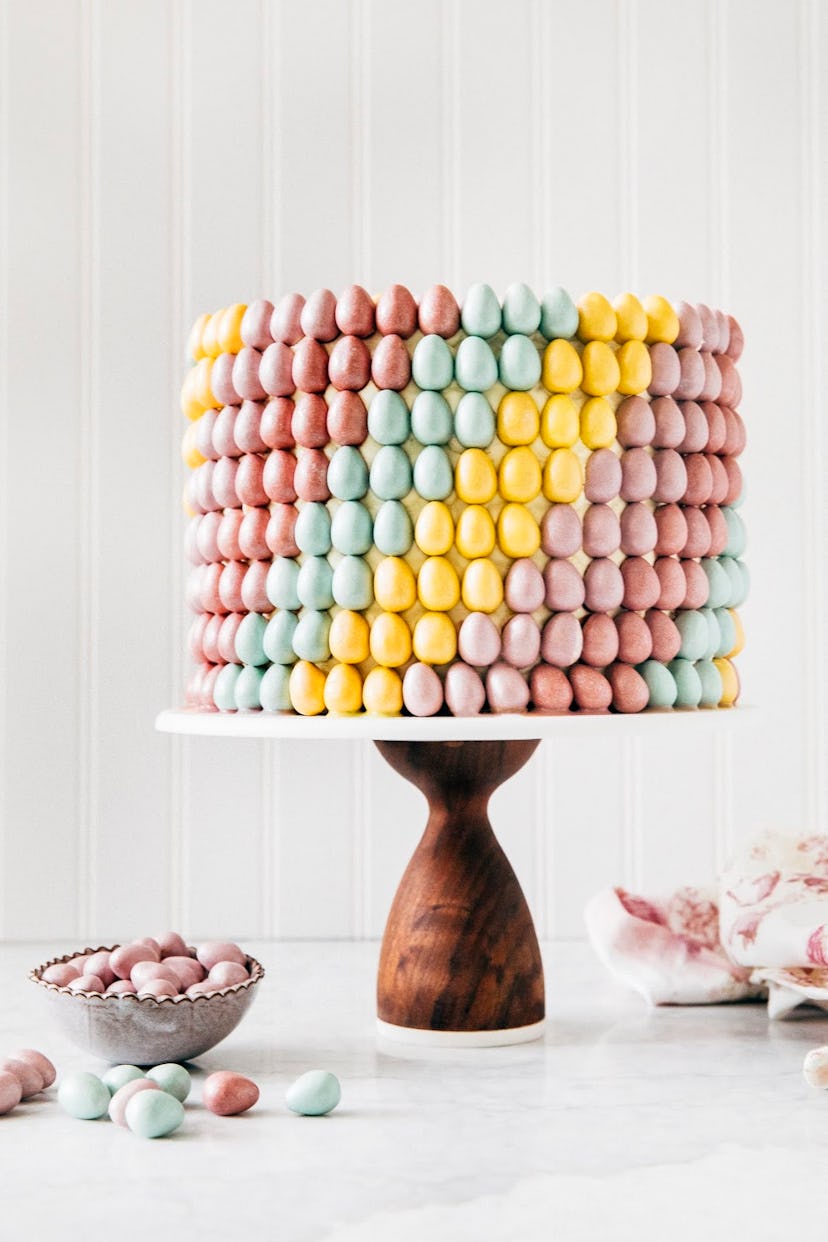 Carrot cake covered in colorful candy eggs in a rainbow swirl pattern on a wooden cake stand with ca...