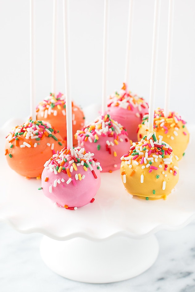 Cake pops covered in pink, orange, and yellow coating with sprinkles displayed on white cake stand