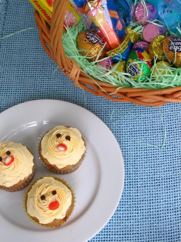 Three cupcakes decorated to look like chicks on a white plate with an Easter basket in the backgroun...