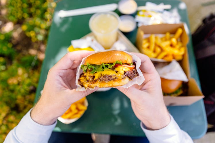 Hands holding a hamburger and a table with a drink and fries