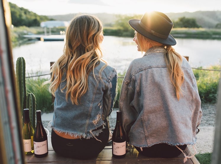 Two best friends sit on the dock at the Trailer Pond at Alta Colina Vineyard with bottles of wine.