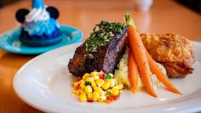 Steak, carrots, mashed potatoes, and corn are on a plate with a colorful Mickey Mouse dessert in the...