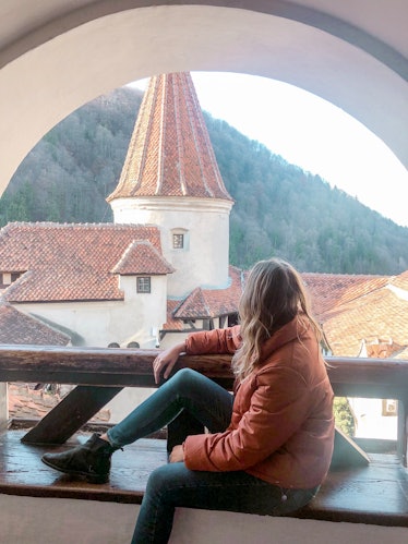 A woman wearing an orange jacket, jeans, and boots sits on a bench overlooking a castle.
