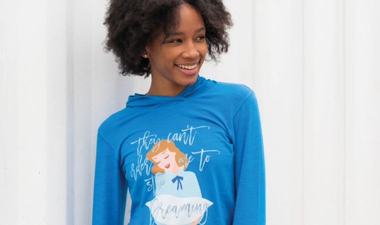 A woman wearing a 'Cinderella' blue hoodie, stands in front of a white wall.