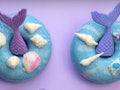 Two mermaid-themed doughnuts are placed on a purple table with seashells. 