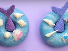 Two mermaid-themed doughnuts are placed on a purple table with seashells. 