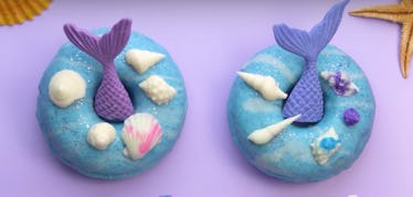 Two mermaid-themed doughnuts are placed on a purple table with seashells. 